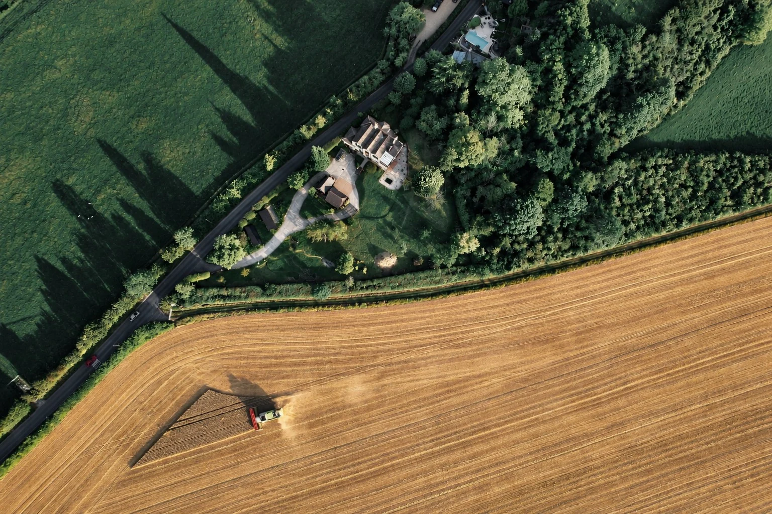 Aerial view of a country house among trees, beside a golden field being harvested